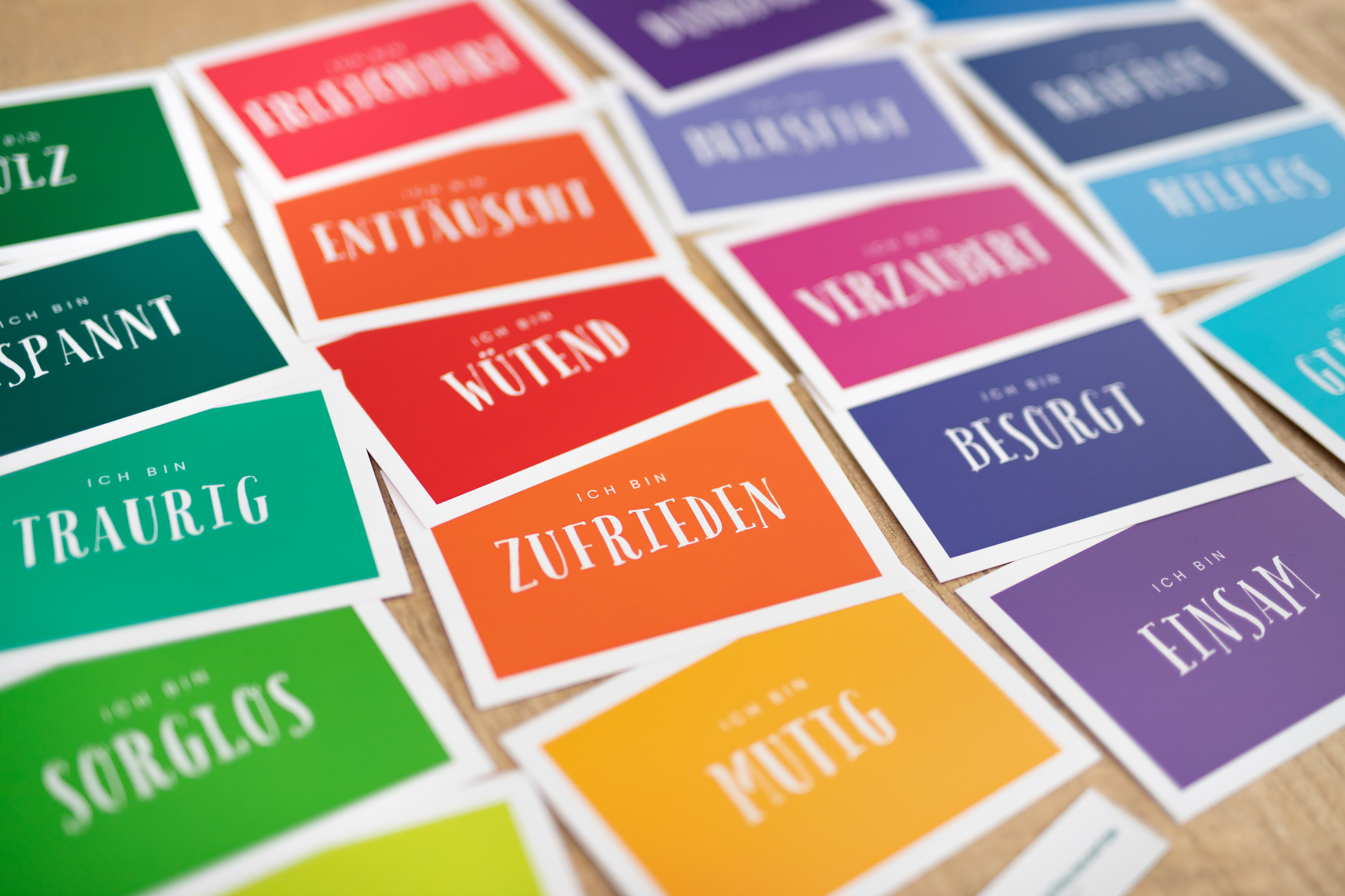 Das Bild zeigt mehrere Gefühlskarten auf denen Emotionen in Form von Wörtern zu sehen sind. Die Karten sind auf einer Tischfläche verteilt. Die Karten sind bunt gestaltet und wirken sehr ansprechend. Zu lesen sind Aufdrucke wie traurig, sorglos, besorgt, zufrieden oder wütend.