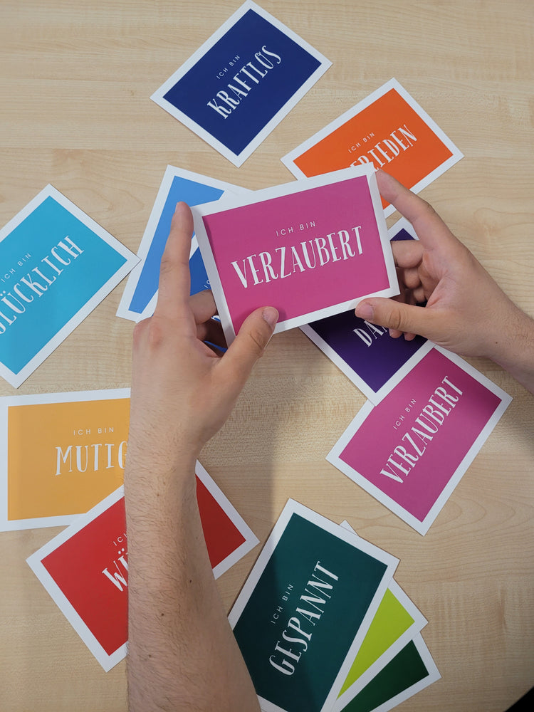Auf dem Foto sind mehrere bunte Karten zu sehen, die alle unterschiedlich mit Gefühlen beschriftet sind. Zwei Hände halten eine Karte in die Kamera auf der "Ich bin verzaubert" geschrieben ist.