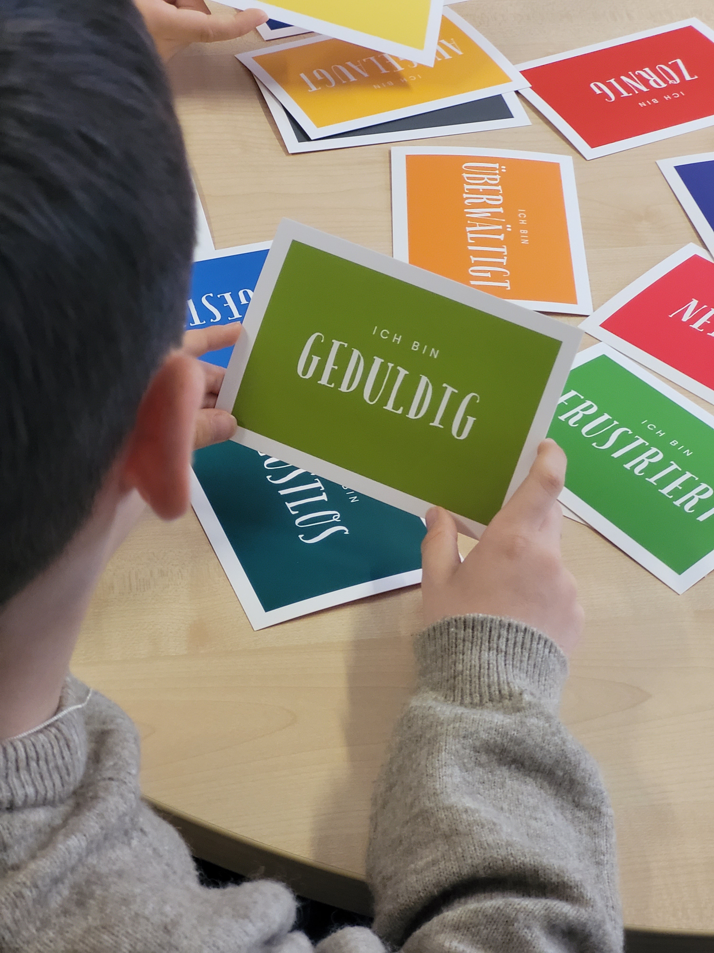 Das Foto zeigt mehrere bunte Karten auf denen Gefühle beschrieben sind. Teilweise sind die Karten überlagert. Ein Junge hält eine Karte mit der Aufschrift "Ich bin geduldig" in die Kamera. Ist selbst aber nur von hinten zu sehen.