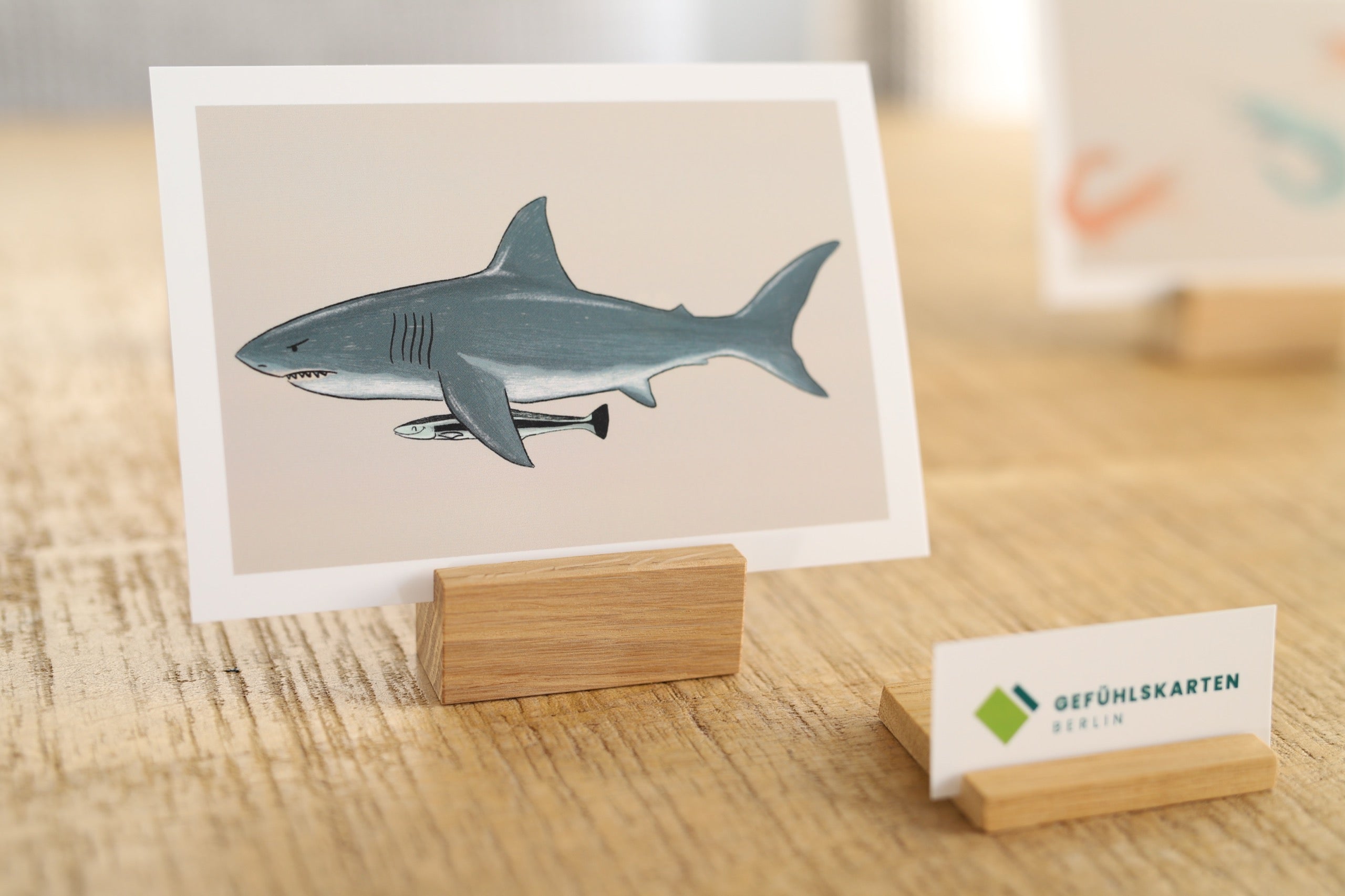 Das Foto zeigt eine Karte in einem Holzkartenhalter. Auf der Karte ist ein Hai und ein Fisch zu sehen. Davor gelagert liegt ein zweiter kleiner Holzkartenhalter, in dem eine Visitenkarten eingesteckt ist.