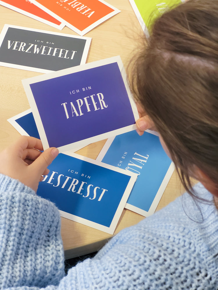 Das Bild zeigt ein junges Mädchen von hinten, welches in ihren Händen eine Karte hält, auf der "ich bin tapfer" zu lesen ist. Etwas verdeckt dahinter sind noch weitere bunte Karten mit Aufschriften. Es handelt sich um die Wortkarten zum Thema Gefühle.