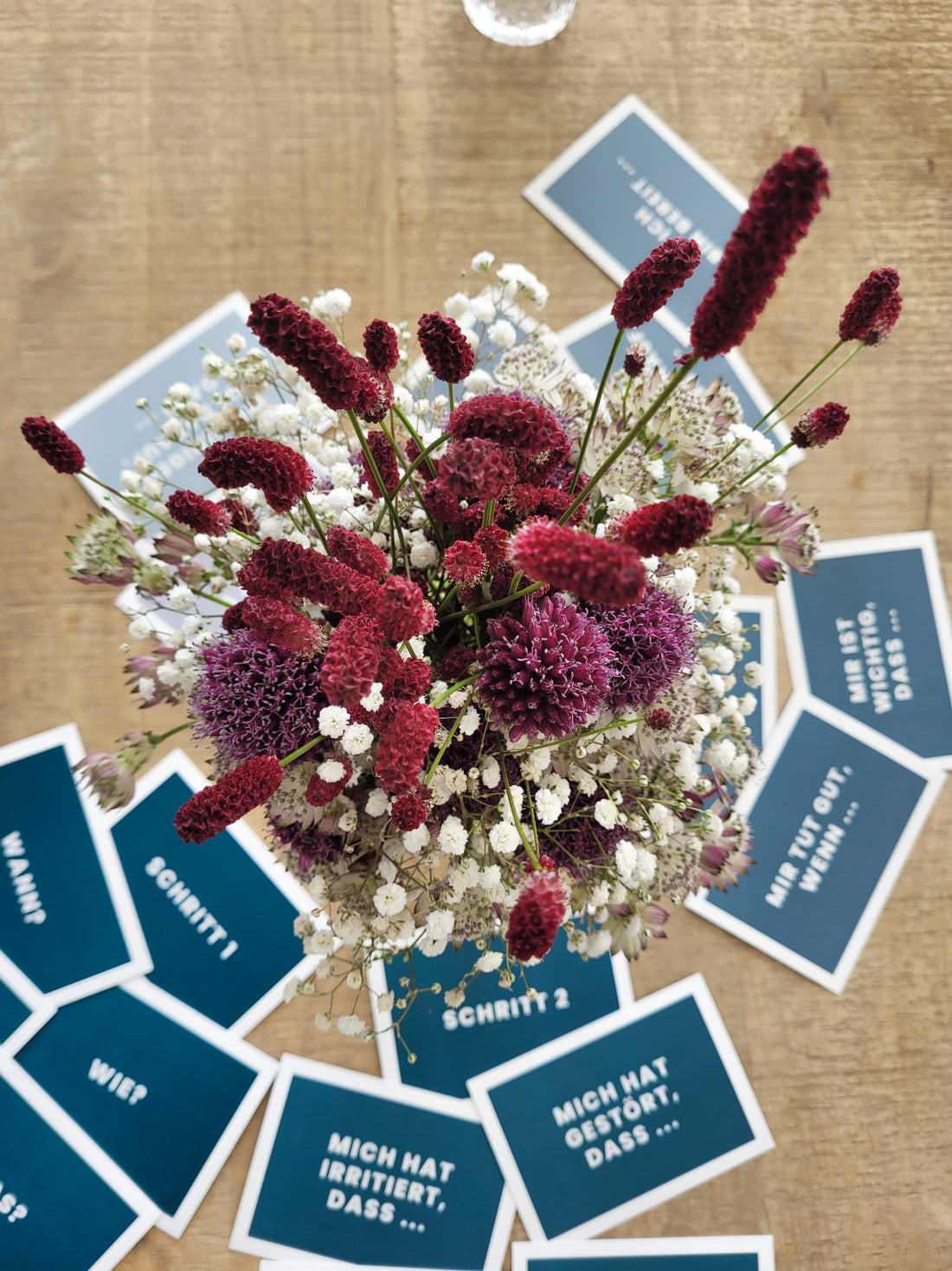 Das Bild zeigt einen Blumenstrauß von oben, um den herum etwas tiefer blau-grau gefärbte Karten mit Schriften liegen. Es handelt sich bei genauerem Hinsehen um die Karten zur Konfliktlösung. 
