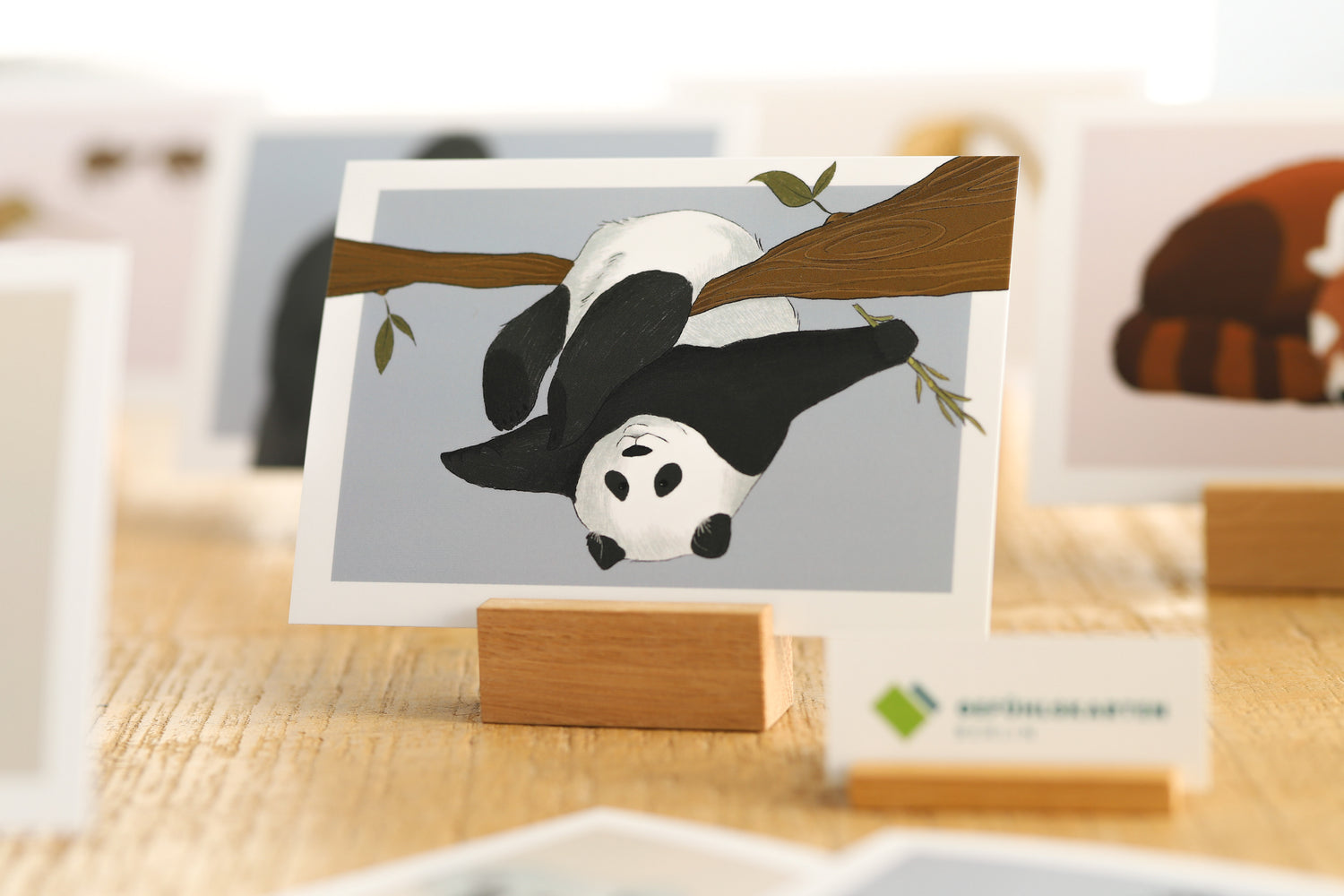 Bild zeigt eine Karte in einem Kartenhalter aus Holz. Auf der Karte ist ein Pandabär zu sehen, der munter von einem Baumast schaut und sich dort im Überschlag mit den Beinen eingehakt hat. Im Hintergrund sind weitere Gefühlskarten mit Tiermotiven verschwommen zu erkennen. 