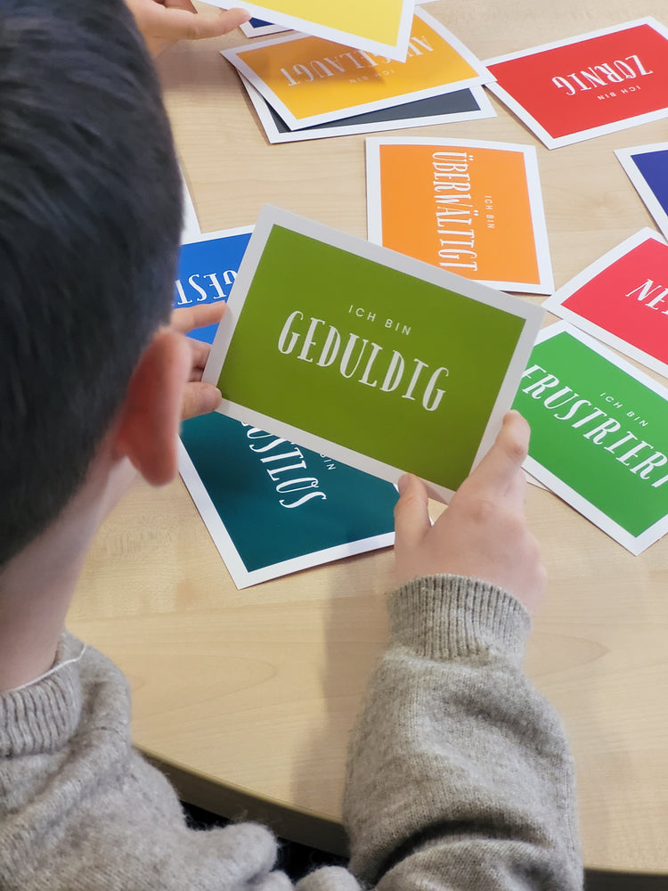 Das Bild zeigt einen Jungen von hinten der  an einem Tisch sitzt und sich mit den bunten Wortkarten zum Thema Gefühle beschäftigt. In der Hand hält er eine ausgewählte Karte mit der Aufschrift: "ich bin geduldig". Auf der Tischfläche sind noch weitere Karten ausgelegt. 