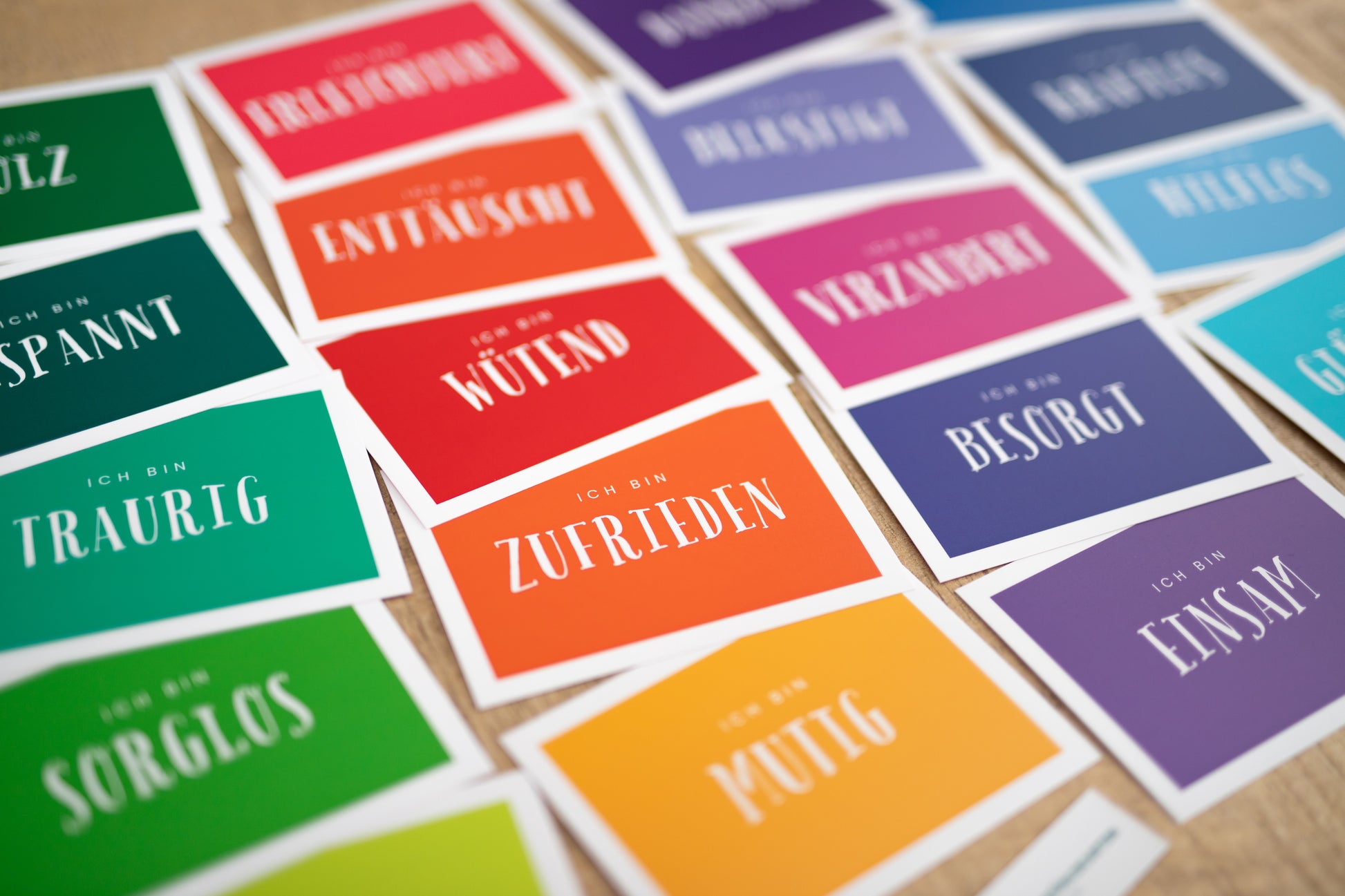 Das Bild zeigt mehrere Gefühlskarten auf denen Emotionen in Form von Wörtern zu sehen sind. Die Karten sind auf einer Tischfläche verteilt. Die Karten sind bunt gestaltet und wirken sehr ansprechend. Zu lesen sind Aufdrucke wie traurig, sorglos, besorgt, zufrieden oder wütend.