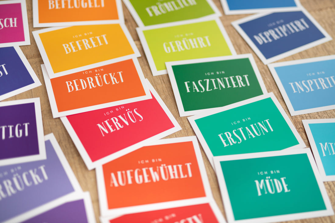 Das Bild zeigt mehrere Gefühlskarten auf denen Emotionen in Form von Wörtern zu sehen sind. Die Karten sind auf einer Tischfläche verteilt. Die Karten sind bunt gestaltet und wirken sehr ansprechend. Zu lesen sind Aufdrucke wie traurig, sorglos, besorgt, zufrieden oder wütend.